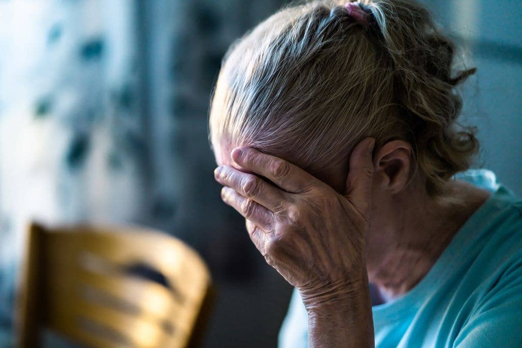 An older woman with short hair covers her face with her hands, showing distress.