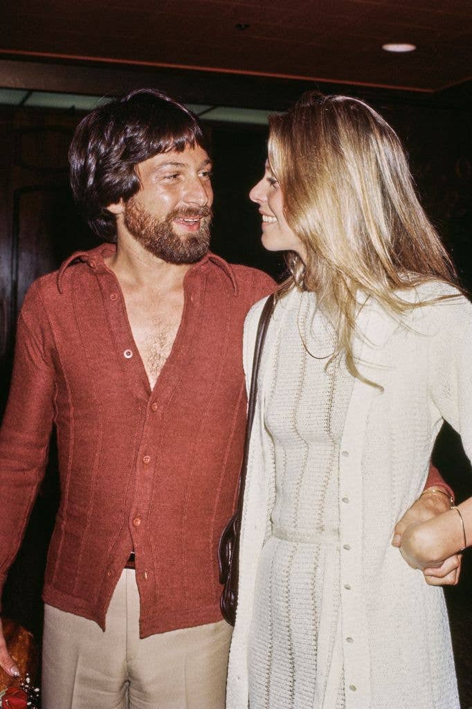 Michael Brandon wearing a red open-collar shirt and white trousers, and his wife, American actress Lindsay Wagner, wearing a long white cardigan over a striped white outfit.