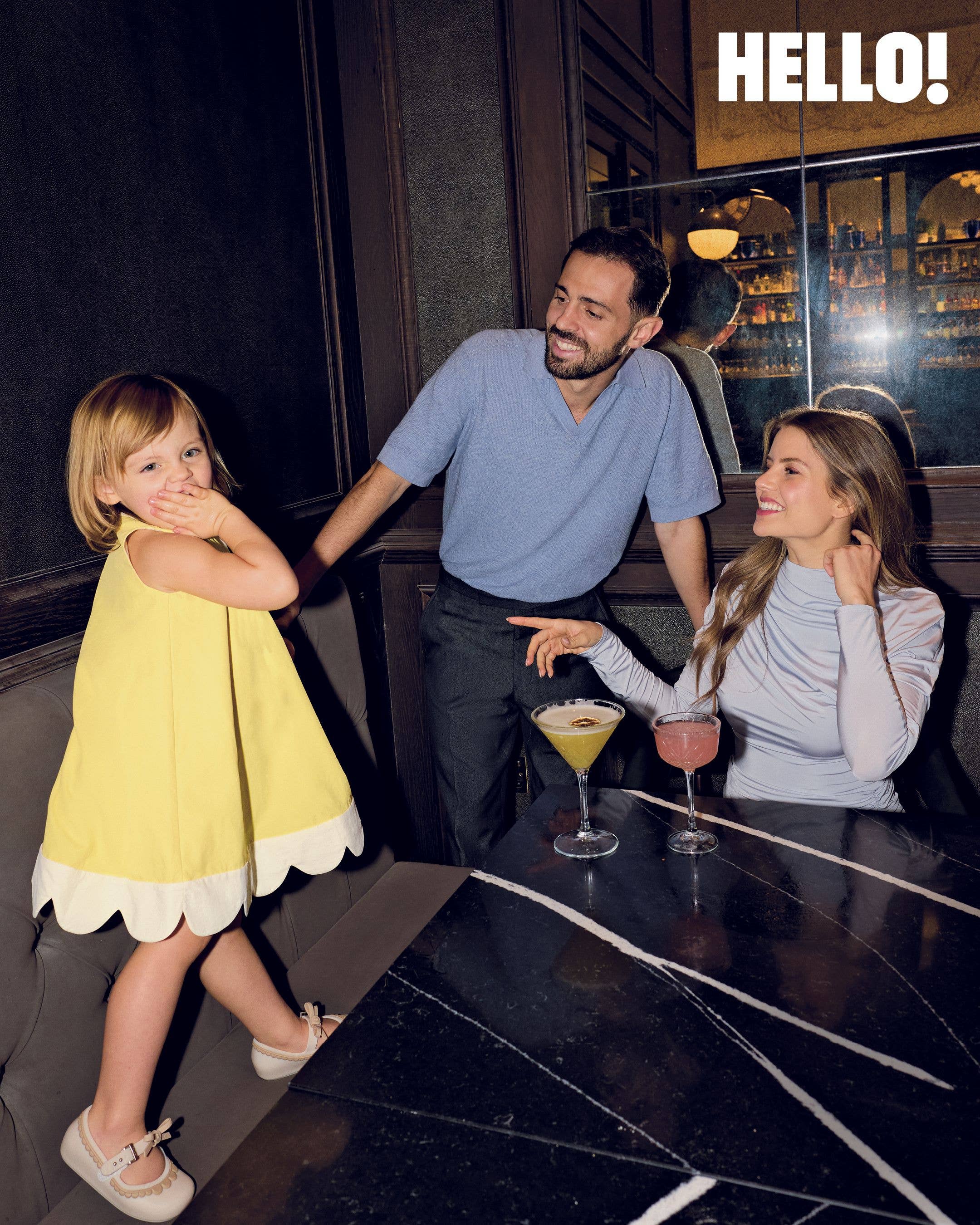 Carlota, wearing a yellow dress, looks at the camera as she moves towards her father Bernardo and mother Ines Silva as they sit at a table with cocktails
