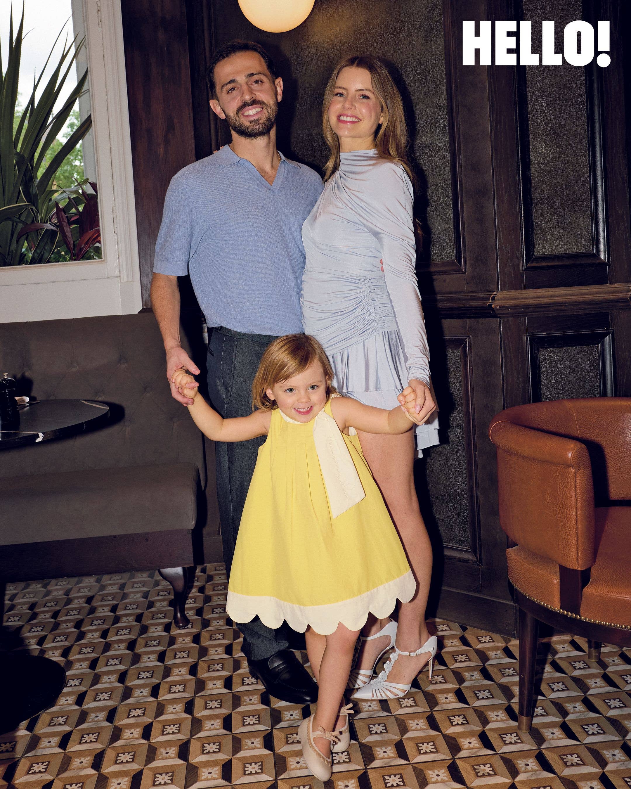 Bernardo and InĂȘs Silva each hold one of their daughter Carlota's hands as she wears a yellow dress with a scalloped hem