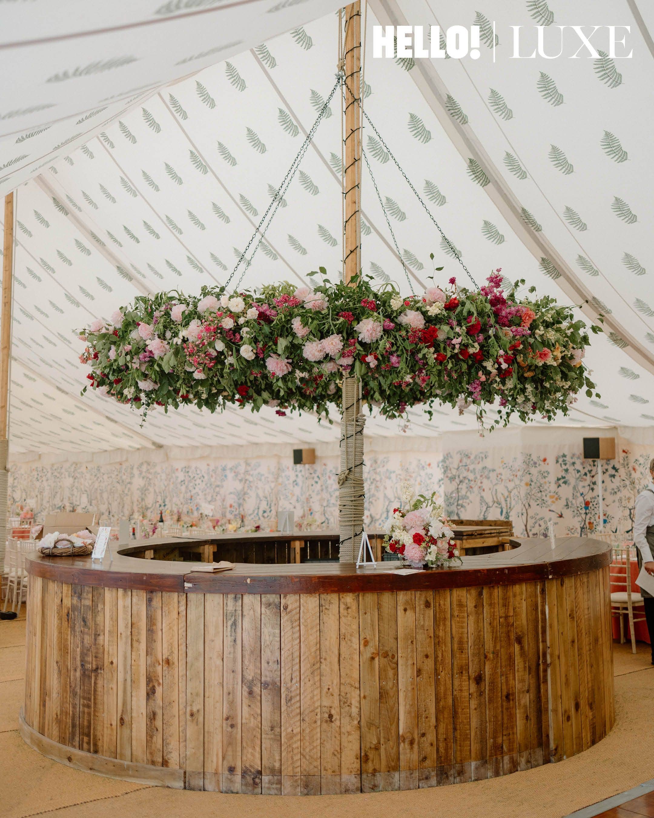The circular wooden bar inside the marquee features a large, elevated floral installation composed of seasonal pink, white, and red summer blossoms