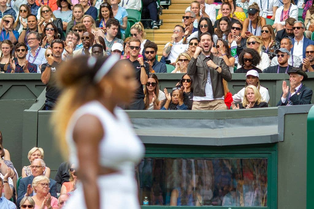 Alexis in focus on his feet in the family box as Serena out of focus in the foreground plays tennis