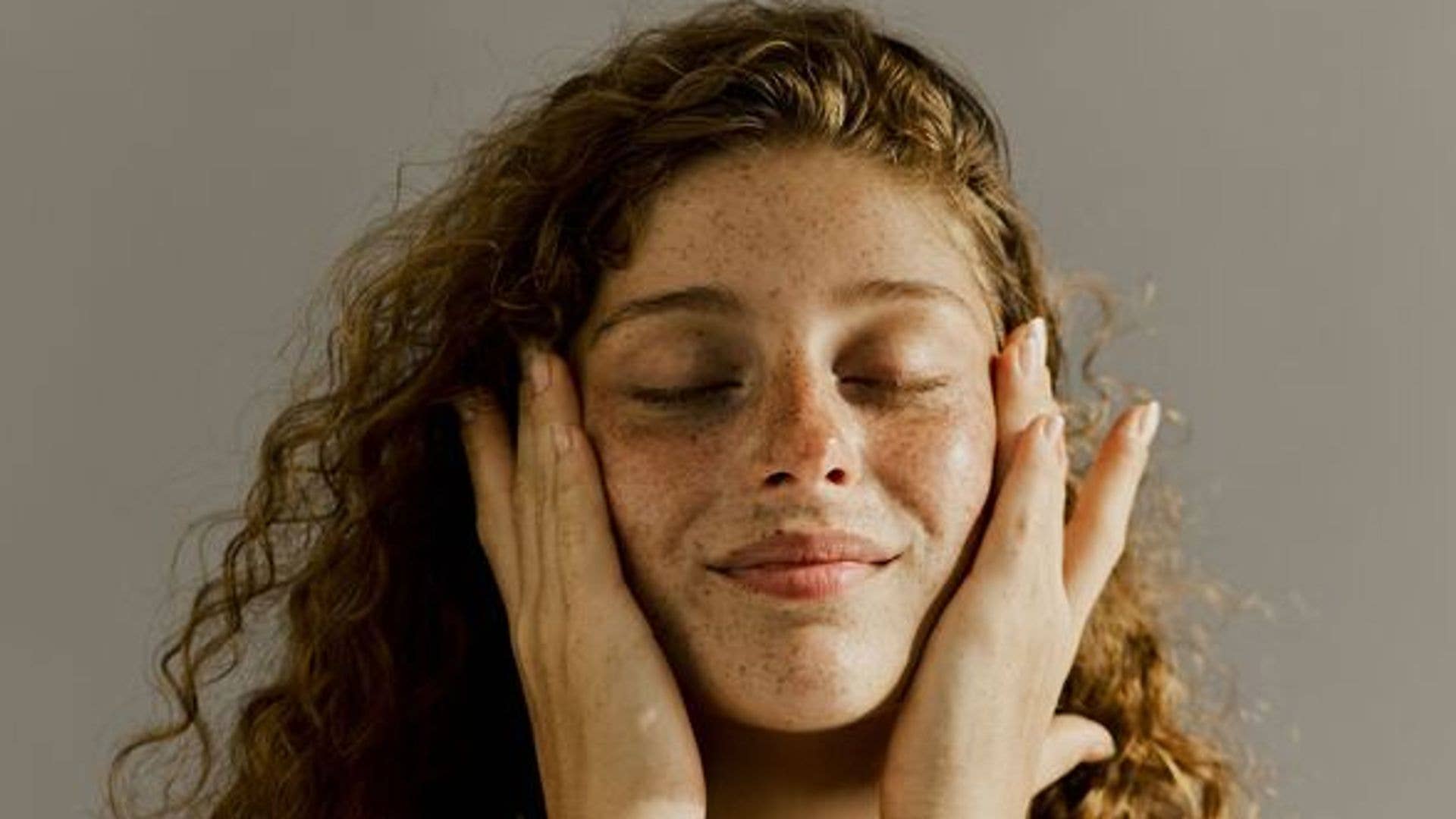 A model with red hair and freckles with her eyes closed holding her face as though she's just applied skincare