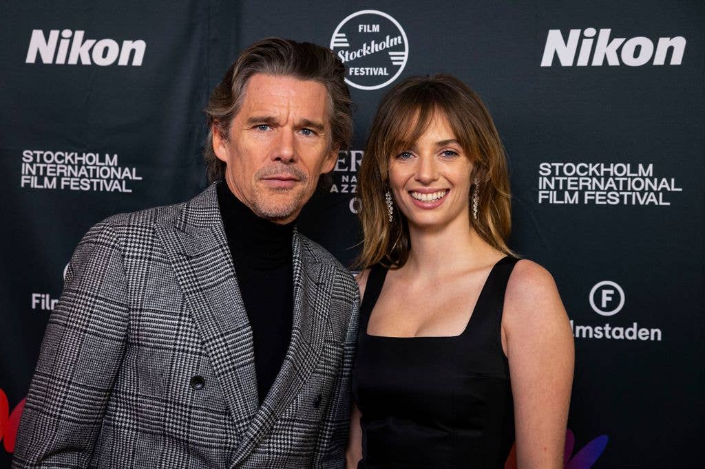 Ethan Hawke and Maya Hawke stand together on the red carpet. Ethan wears a black turtleneck and check grey jacket while Maya wears a strapped black, square neck dress.