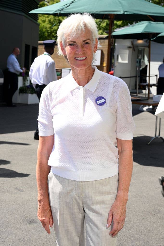 Judy Murray stands smiling in a white polo shirt and khaki pants, badge on her chest.