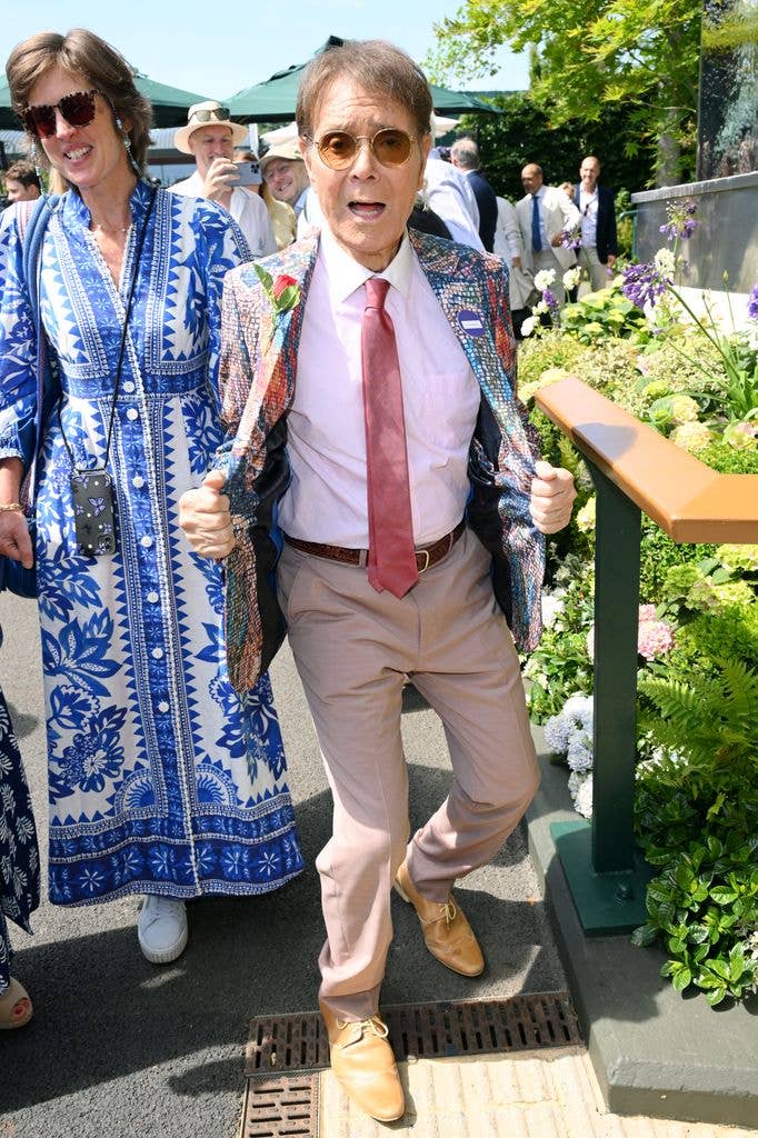 Cliff Richard, wearing a colorful jacket and red tie, walks with a woman in a blue dress.