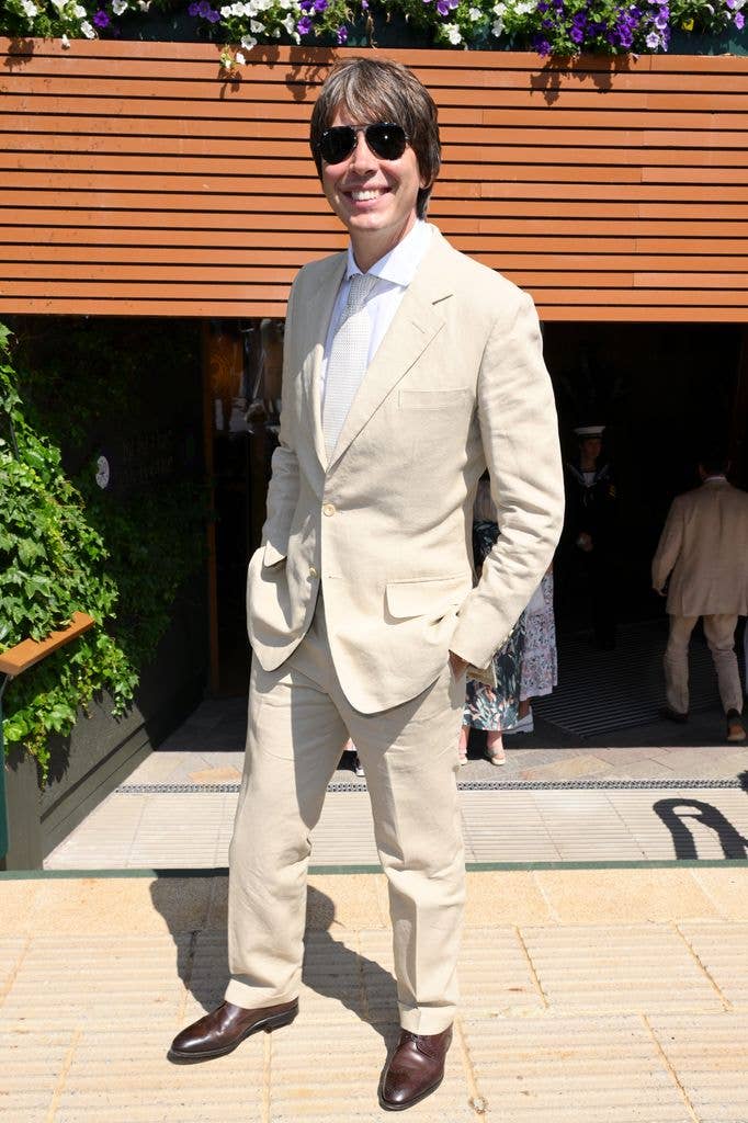 Brian Cox stands smiling in a light beige suit, white shirt, and brown shoes, with hands in pockets.