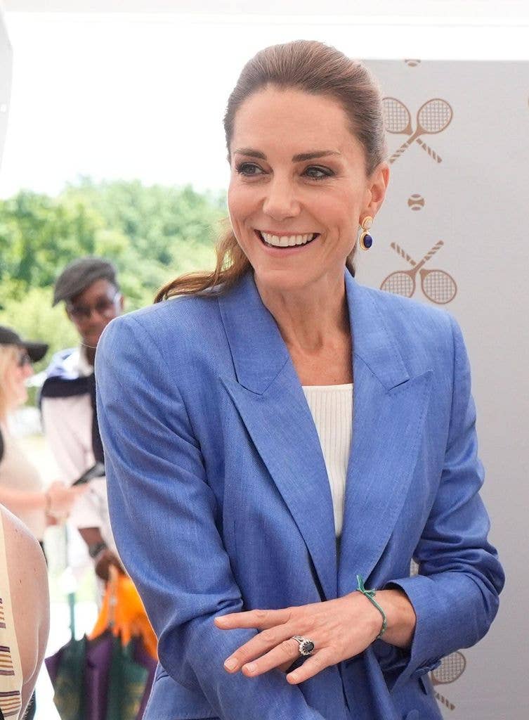 Kate Middleton smiling, wearing a blue blazer and white top, stands in front of a tennis-themed backdrop.
