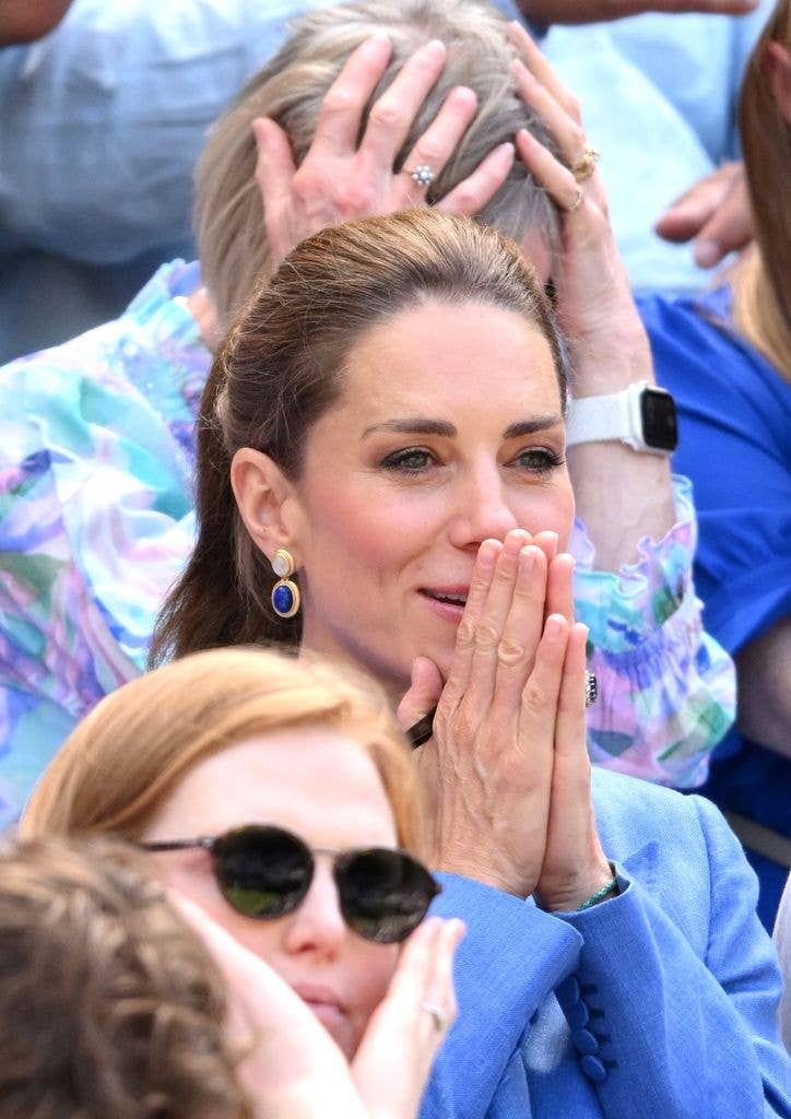 Kate Middleton, wearing blue suit and earrings, claps with hands together at an event.