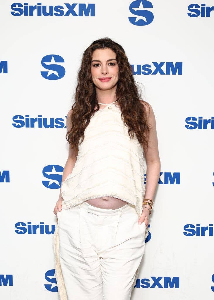 Anne Hathaway stands in front of a Sirius XM backdrop, wearing white attire.