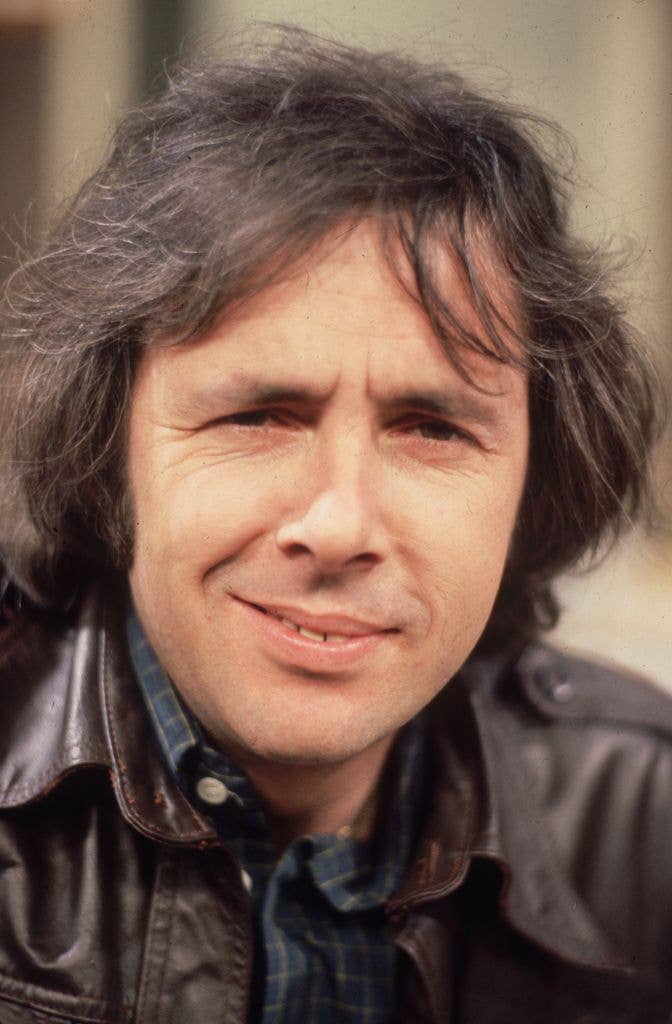A smiling man with long hair wearing a leather jacket and plaid shirt.