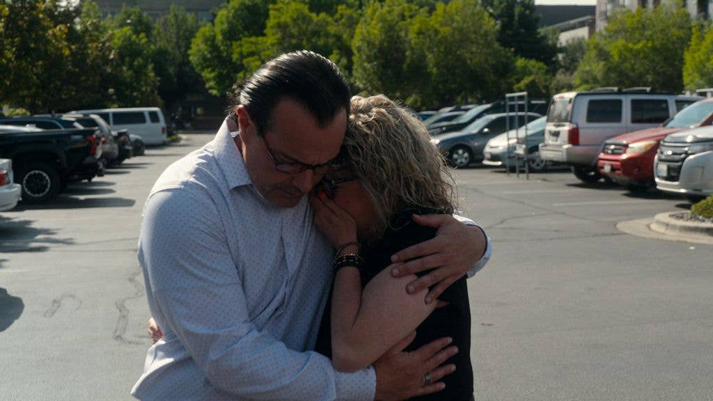 A man and a woman embrace in a parking lot, both wearing casual clothing.
