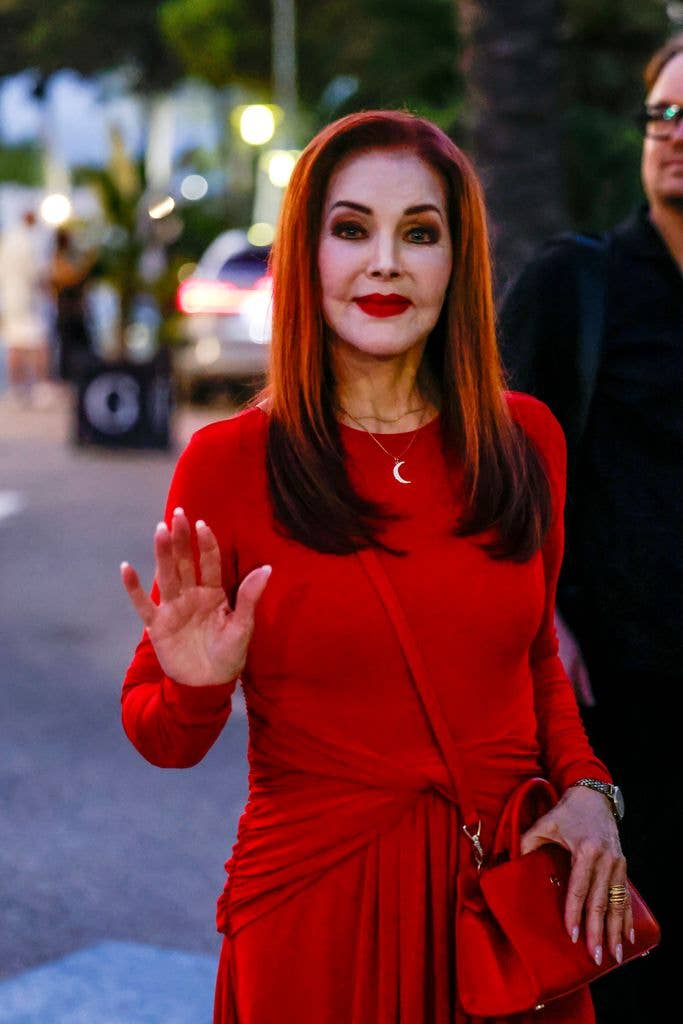 Priscilla Presley waves while wearing a red dress and holding a red purse.