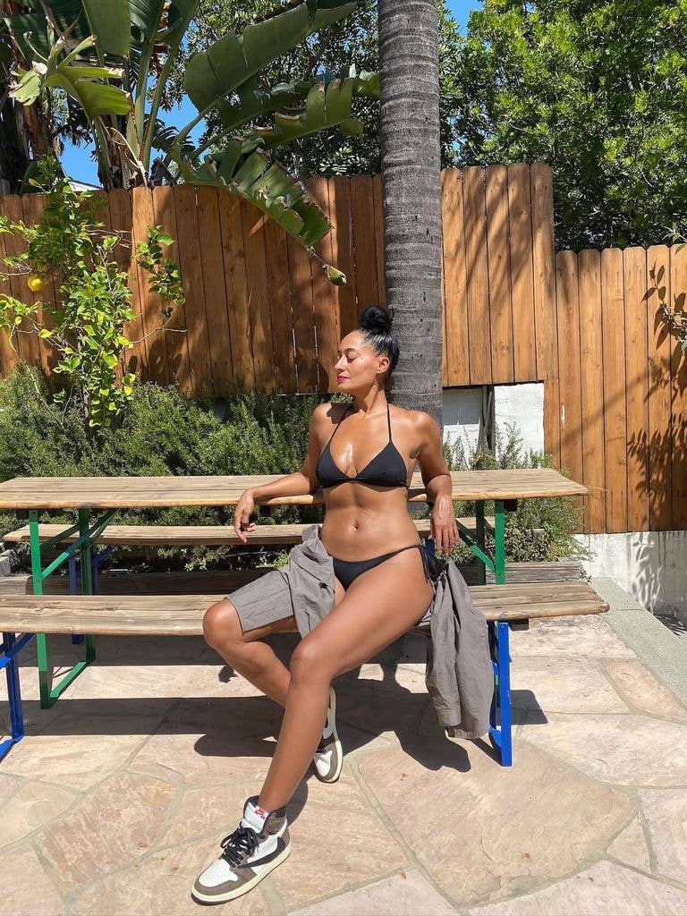 Tracee Ellis Ross sitting on a picnic table in a black bikini and gray shorts.