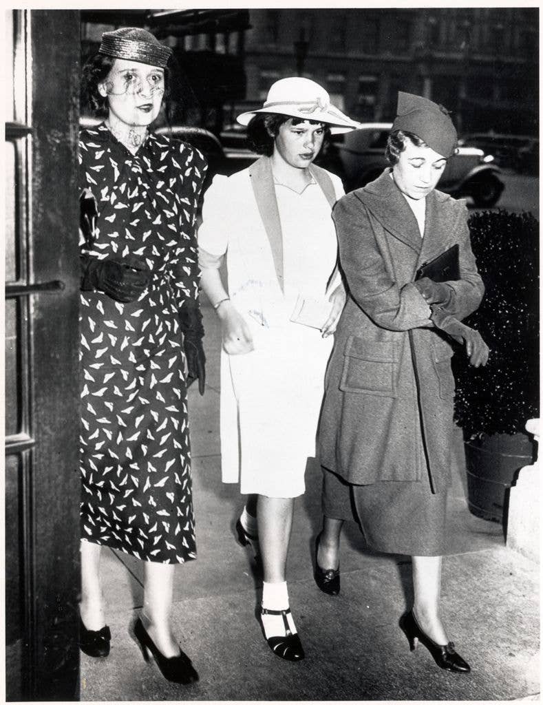 Three women in vintage attire, one in a patterned dress, another in a white suit, and the third in a long coat, walking together.