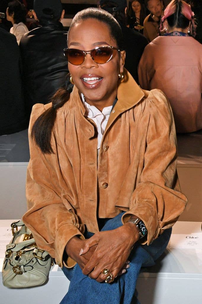 Oprah Winfrey wearing sunglasses and a brown jacket, smiling at a fashion event.
