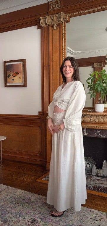 Anne Hathaway stands in front of a mirror, smiling, wearing a white robe.