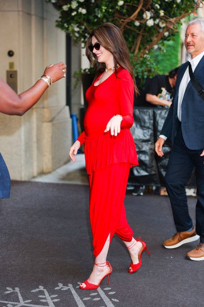 Anne Hathaway in red dress and sunglasses, walking and smiling, with two people nearby.