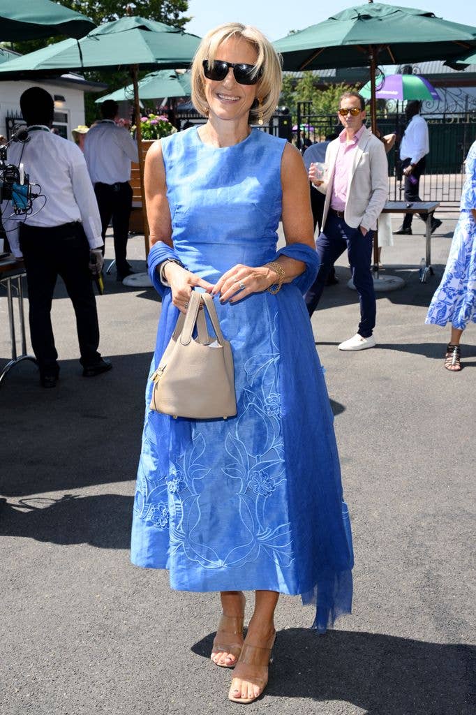 Emily Maitlis in a blue dress and sunglasses, holding a beige handbag, stands smiling at an outdoor event.