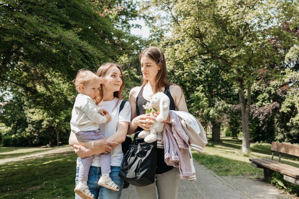 Two women and a child stand in a park; one woman holds a stuffed toy and jackets.