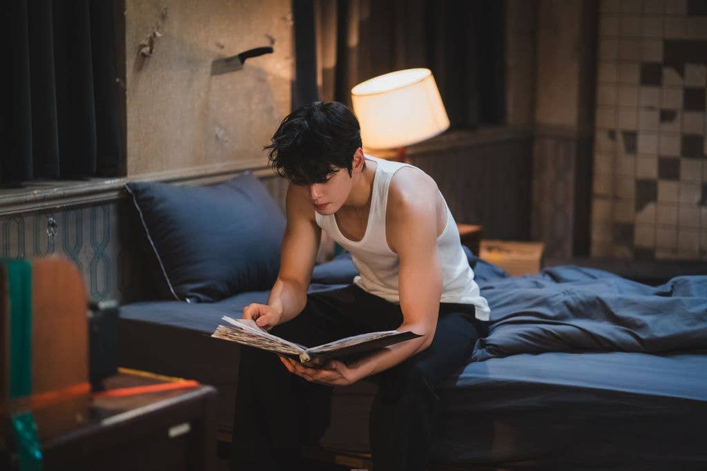 Cha Eun-woo reading a book while sitting on a bed in a dimly lit room.