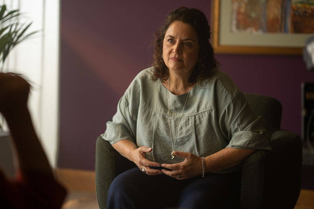 Ruth Jones sits on a chair, hands clasped, wearing a green blouse and necklace.