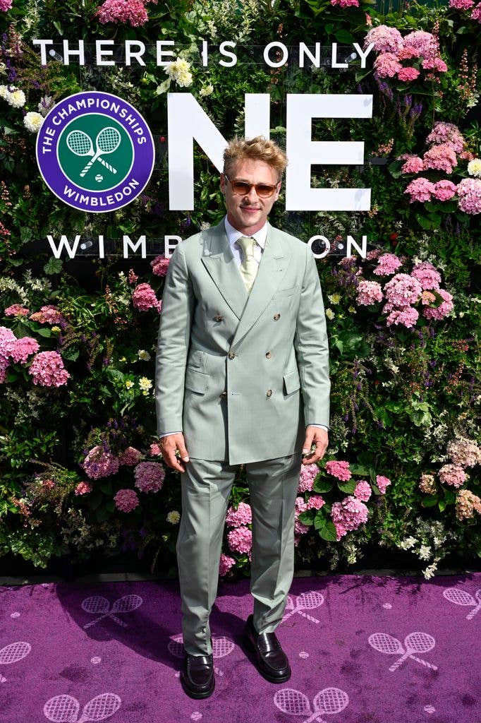 Ben Hardy stands on a purple carpet with tennis racket designs, wearing a light green suit and sunglasses, in front of a floral backdrop.