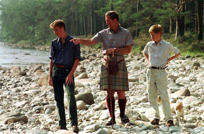 Charles, Prince of Wales, points to something while standing with two young men on rocky ground near a river.