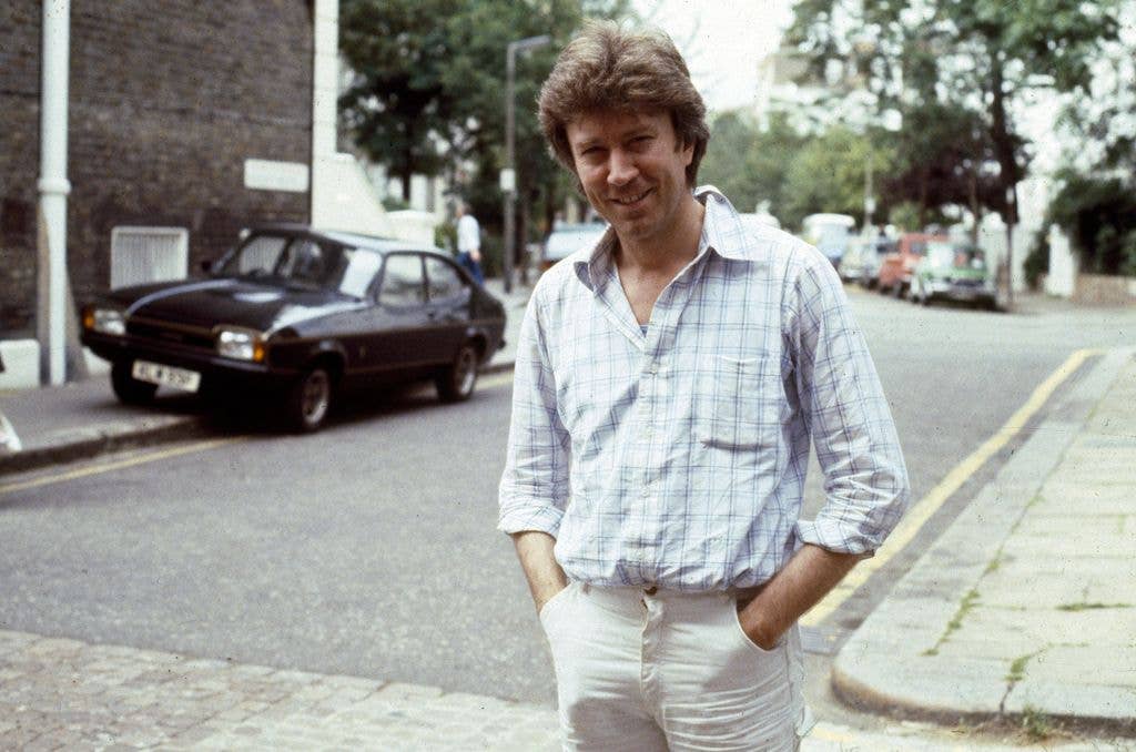Robin Ellis stands on a street with hands in pockets, wearing a plaid shirt and white pants.