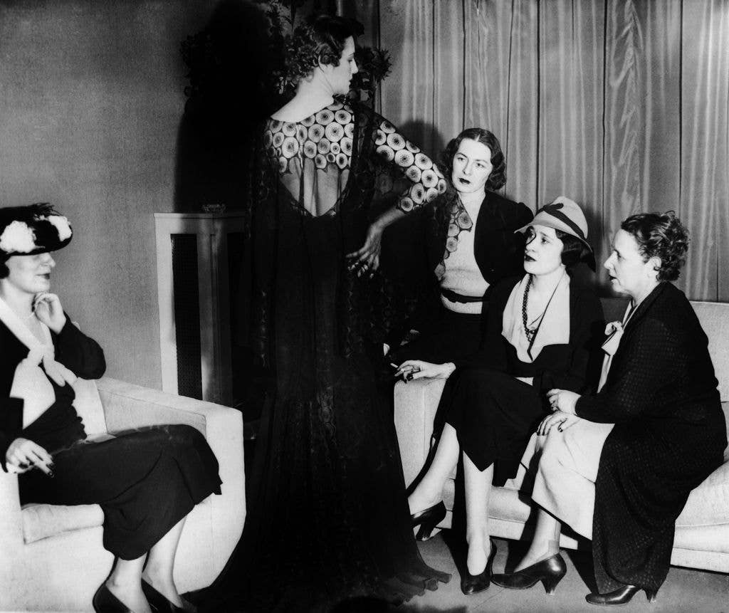 Five women, including Gloria Morgan Vanderbilt and Vicountess Furness, in 1930s-era clothing, one standing, four seated, engaged in conversation
