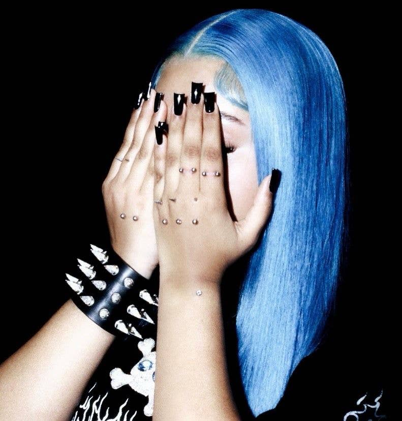North West with blue hair, black nails, and spiked wristband covering their face with hands.