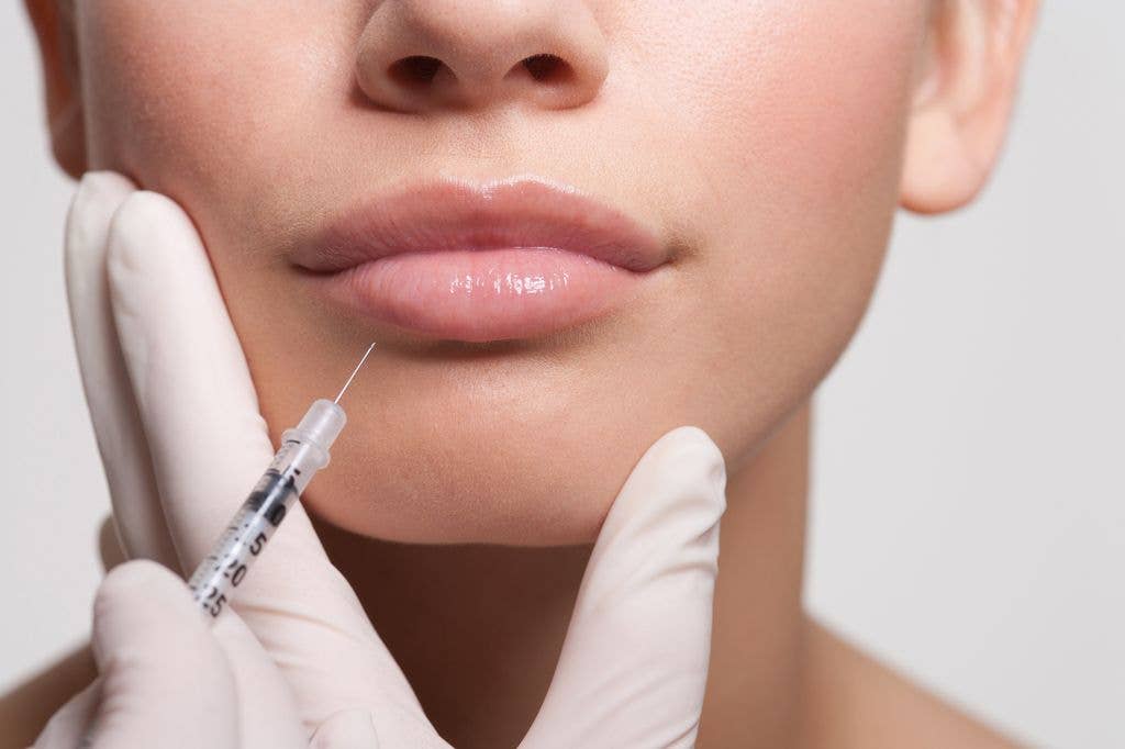 A  woman is receiving a lip injection with a syringe held by gloved hands