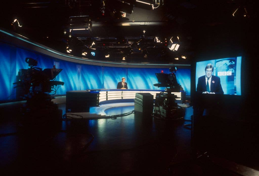 News studio with a man in a suit and tie at a desk, and another man on a BBC screen.