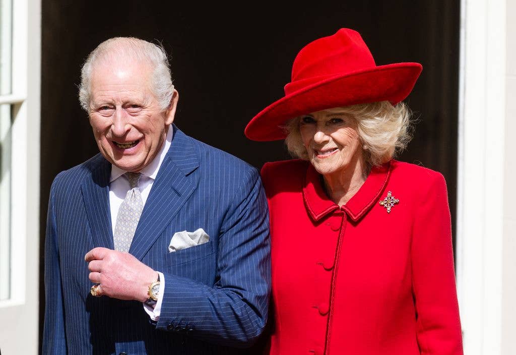 King Charles in a blue suit stands next to Queen Camilla in a red coat and red hat, both smiling.