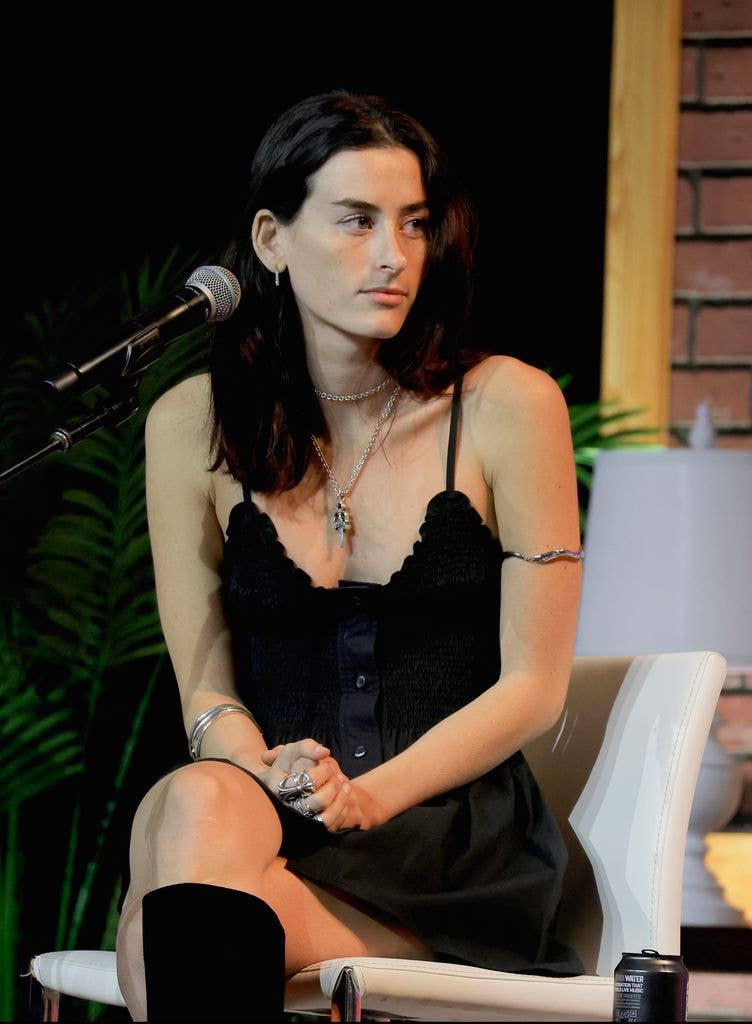 Stella Lefty in a black dress sits on a white chair, facing a microphone.