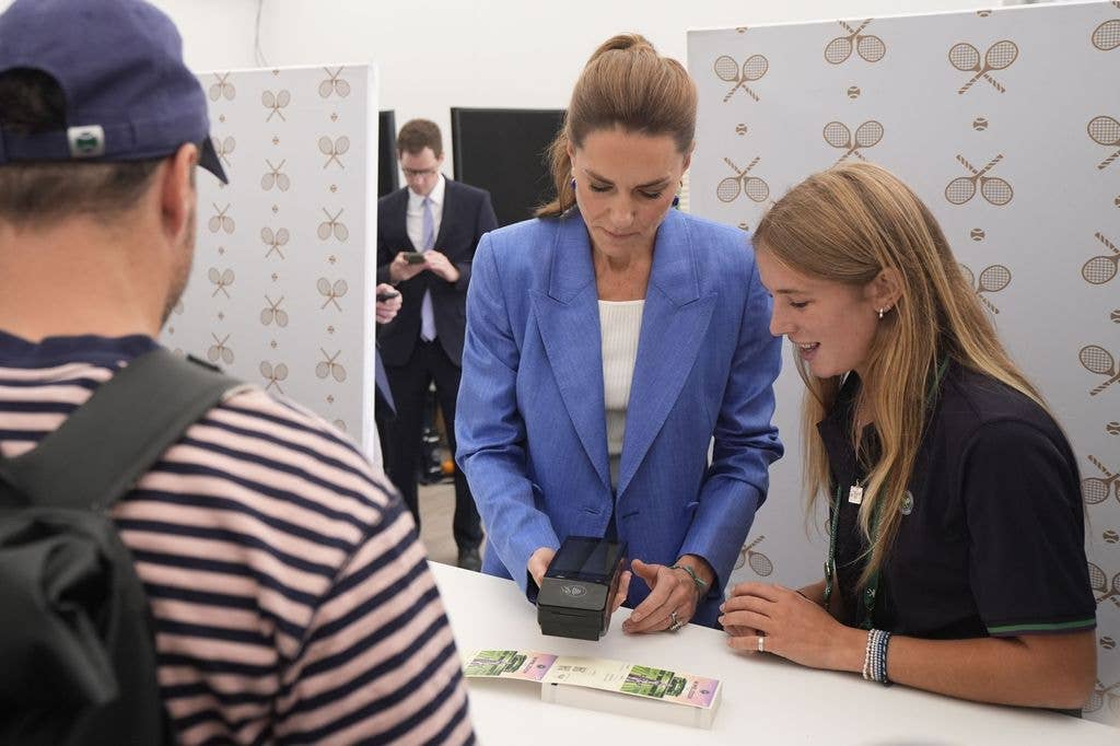 Kate Middleton in a blue blazer scans a ticket while a smiling woman in a black shirt watches