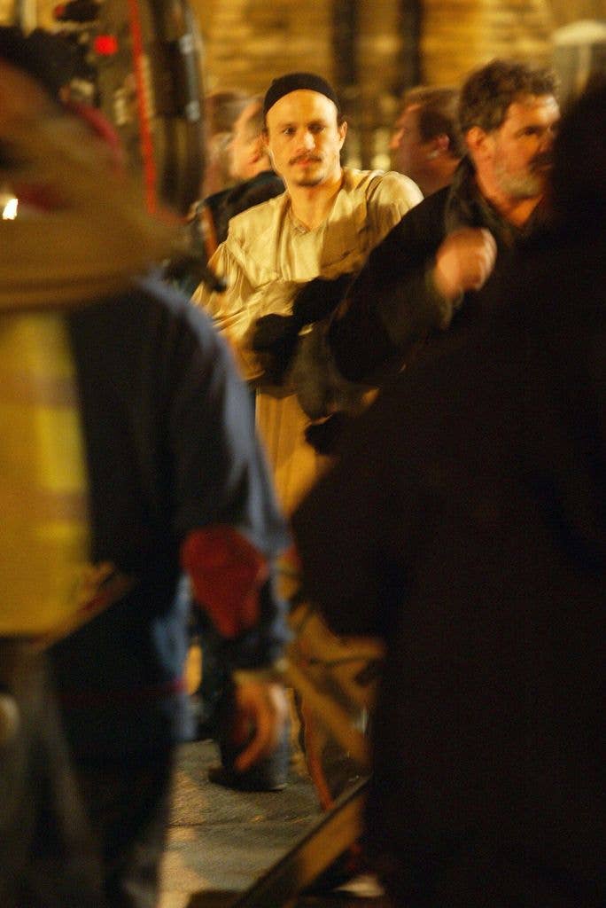 Heath Ledger, wearing a beige shirt and black hat, stands among a crowd.