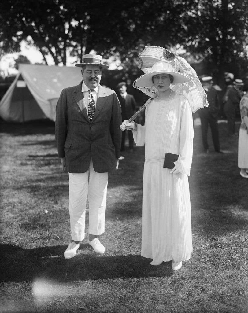 Mr. Reginald Vanderbuilt and Miss Gloria Morgan in vintage attire in 1923, he in suit and hat, she in dress and hat, holding an umbrella