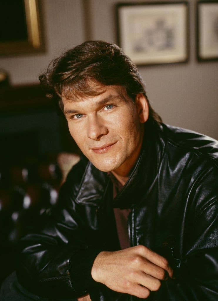 Patrick Swayze in a leather jacket with his arms crossed on top of a table. 