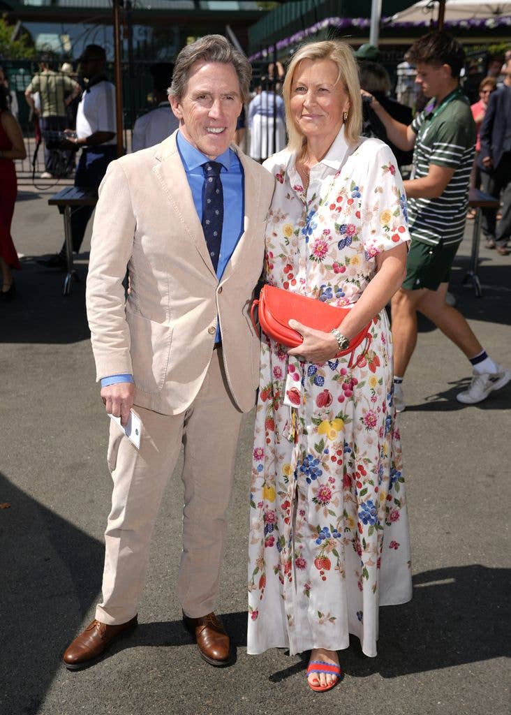 Rob Brydon stands next to a woman in a floral dress at an outdoor event.