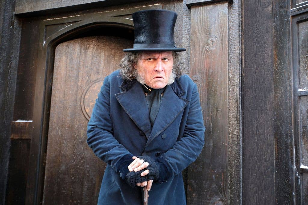 A man in a blue coat and top hat stands by a wooden door, holding a cane.