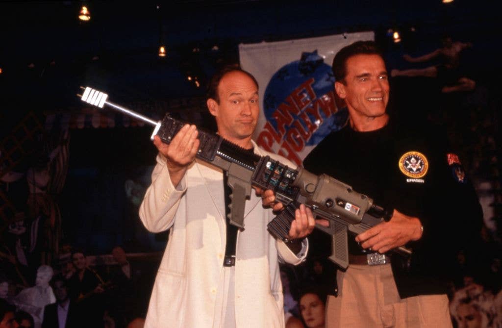 Arnold Schwarzenegger and Chuck Russell holding a large gun, smiling at an event.