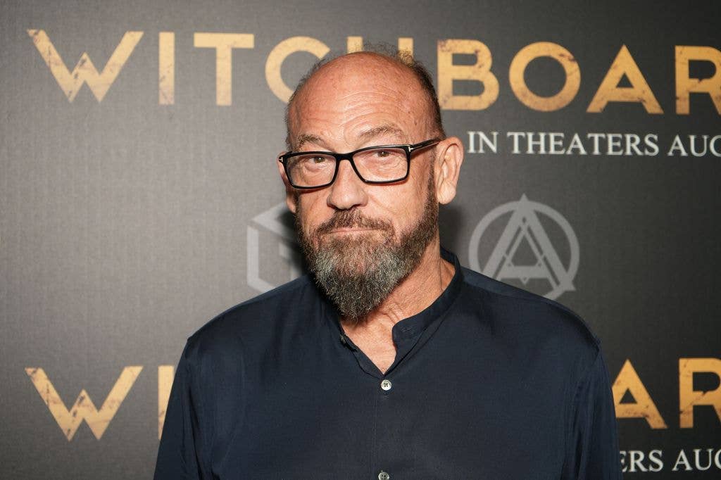Chuck Russell, with a beard and glasses, stands in front of a 'Witchboard' movie poster.