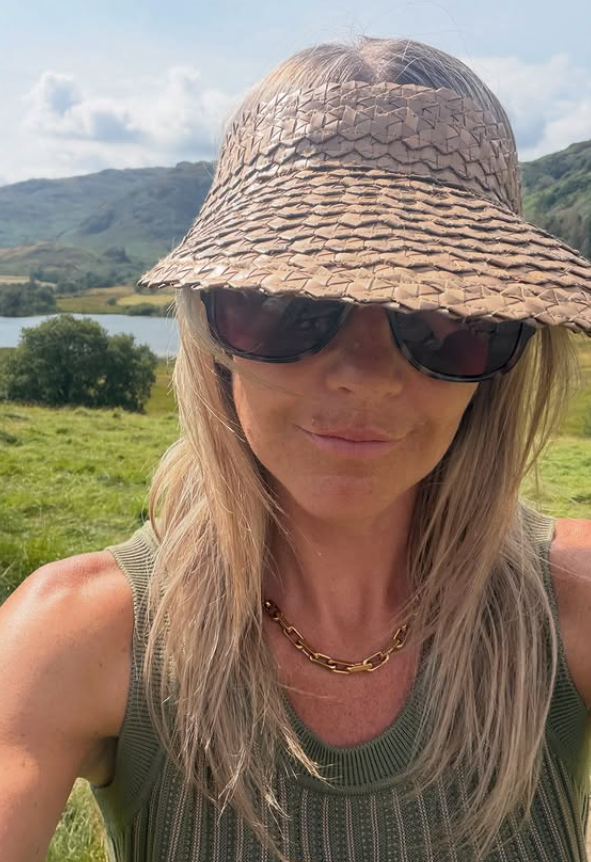 Woman with long blonde hair wearing a straw hat and sunglasses in a grassy field.