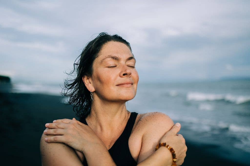 Woman with short dark hair, wearing a black tank top and a beaded bracelet, standing on a beach with eyes closed and arms crossed.
