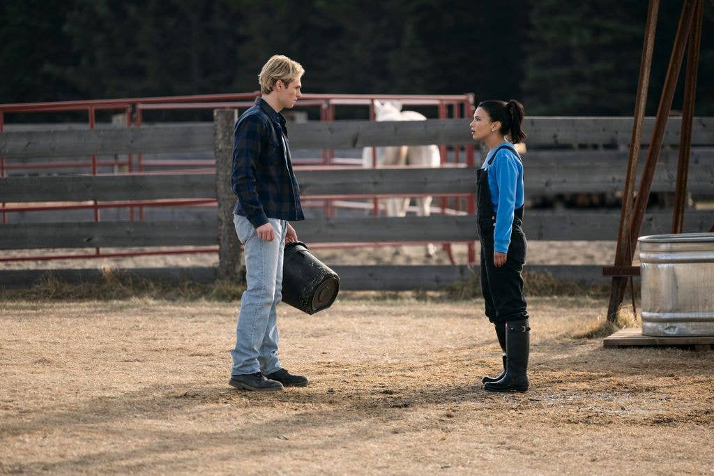 Man in plaid shirt and jeans holds a bucket, facing woman in blue shirt and black overalls in a farm setting.