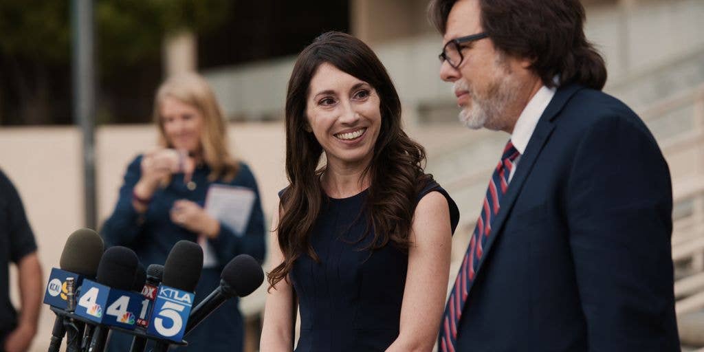A woman in a black dress stands next to a man in a suit and red tie, both facing microphones.