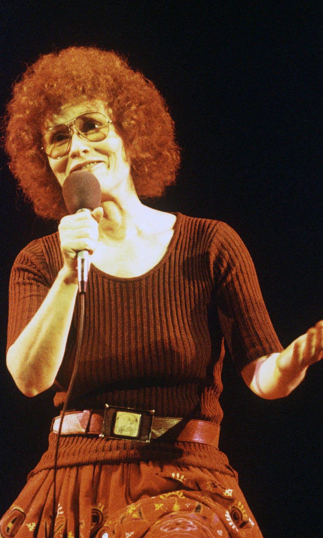 Dory Previn, with red curly hair and glasses, holds a microphone on stage.