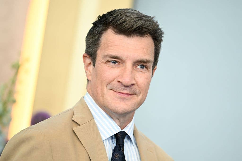 Nathan Fillion smiling, wearing a beige jacket and blue striped shirt with a black tie.