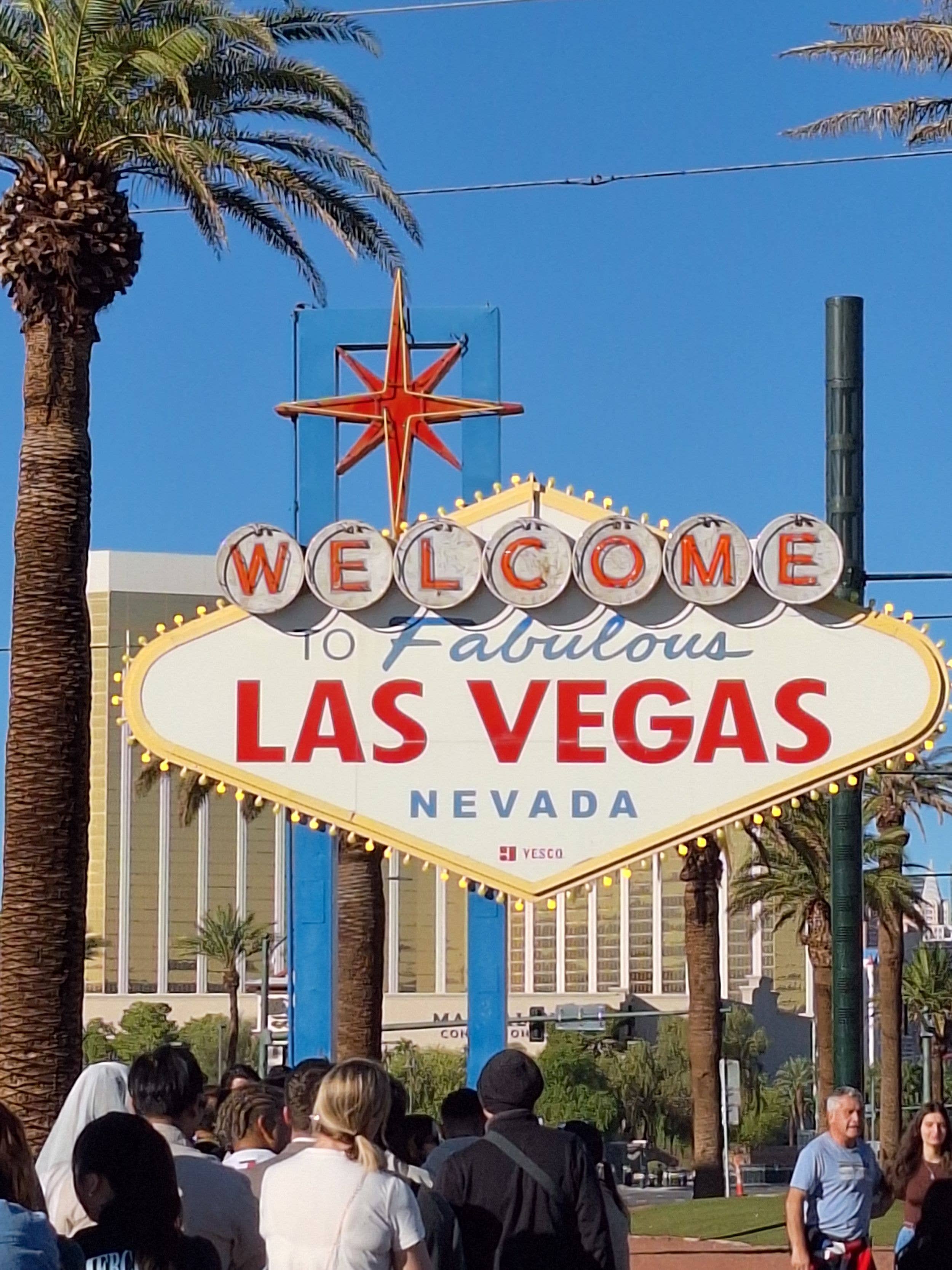 Crowd of people under the iconic 'Welcome to Fabulous Las Vegas' sign with palm trees and buildings.